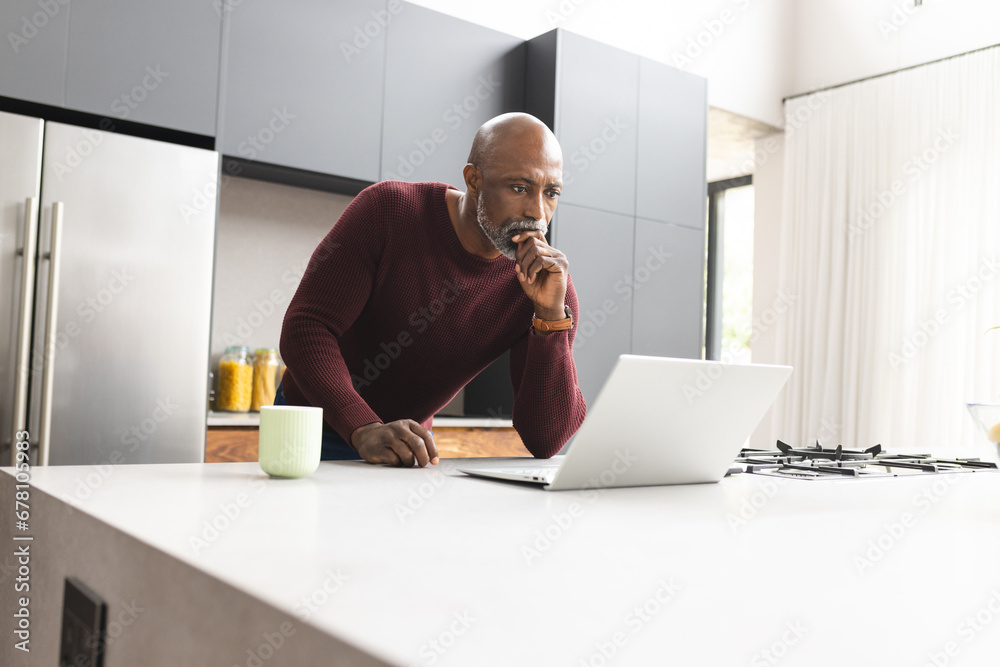 Focused mature african american man having coffee using laptop standing in sunny kitchen, copy space Focused mature african american man having coffee using laptop standing in sunny kitchen, copy space