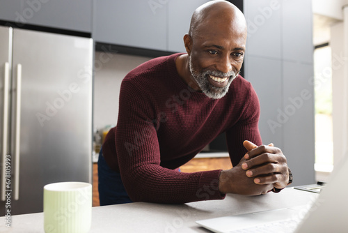 Happy mature african american man having coffee using laptop standing in sunny kitchen photo