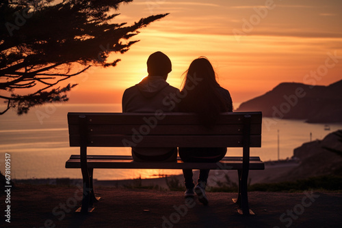 Rear back lit view of unrecognizable young couple sitting on a bench watching sunset over ocean, aesthetic look
