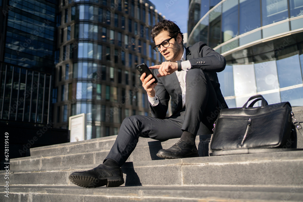 A businessman with glasses in a suit uses a phone and a business app. A successful confident man reads a message on the phone with a colleague at a startup and a new project at work. A businessman with glasses in a suit uses a phone and a business app. A successful confident man reads a message on the phone with a colleague at a startup and a new project at work.