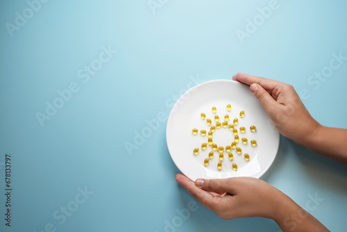Composition with female hands holding a white plate with tablets on inside a picture of the sun on blue background. Creative composition vitamin D. Flat lay space for text.