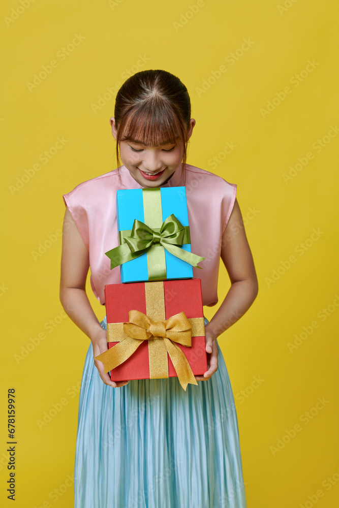 Attractive young girl in dress holding stack of gift boxes isolated over yellow background Attractive young girl in dress holding stack of gift boxes isolated over yellow background