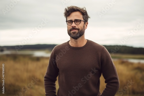 Portrait of a jovial man in his 30s wearing a cozy sweater against a quiet countryside landscape. AI Generation