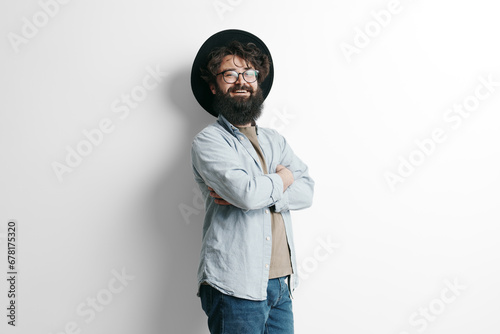 Confident bearded man with glasses smiling and standing arms crossed, wearing a stylish hat and casual shirt, against a white background