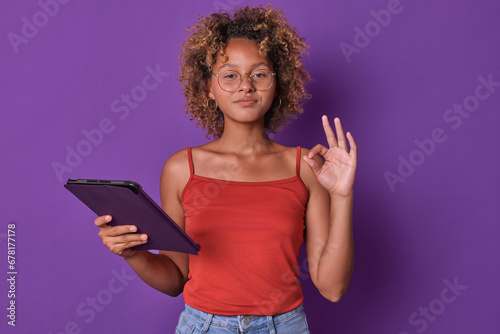 Young successful casual African American woman freelancer holding tablet computer and showing OK gesture after completing customer task or successfully searching for new job stands in purple studio.