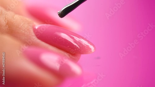 Applying Nail polish, pink shellac UV gel, varnish, manicure process concept in beauty salon. Transparent top coat drop on brush. Over pink background. Application of nail polish close up