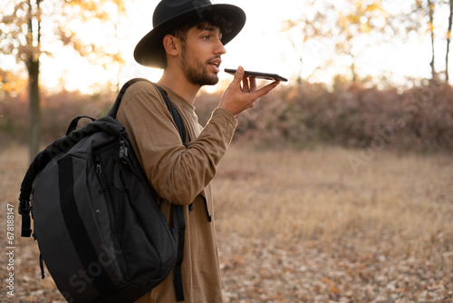 Arabic young man talking on speakerphone, dictating voice message, using online translator app while travel on autumn forest