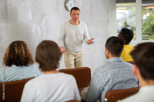 Middle-aged man teacher reading lecture in front of group of guys attending him sitting on chairs
