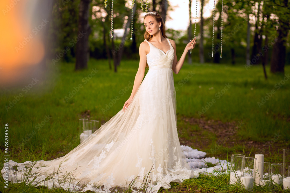 Full-length portrait of a beautiful young slim sexy caucasian bride in fashionable trendy white gown standing on on a decorated outside venue with candles and chandeliers