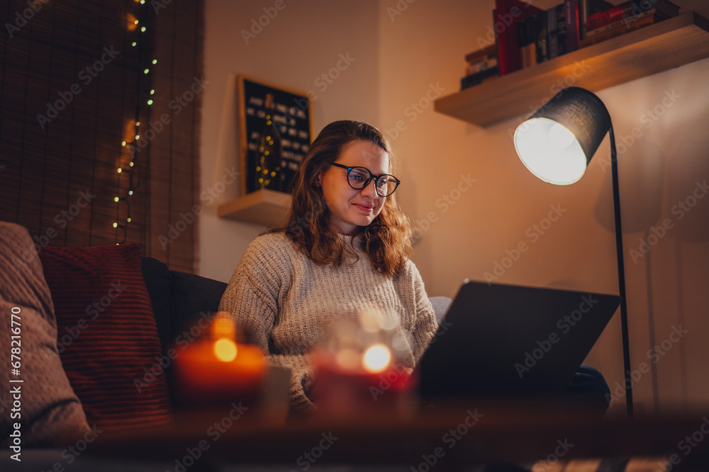 Obraz premium A young woman spends a cozy winter evening at home lying on the sofa with a laptop. Winter holidays, Christmas and online surfing