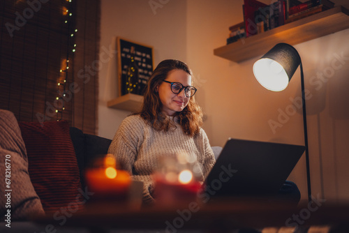 A young woman spends a cozy winter evening at home lying on the sofa with a laptop. Winter holidays, Christmas and online surfing photo