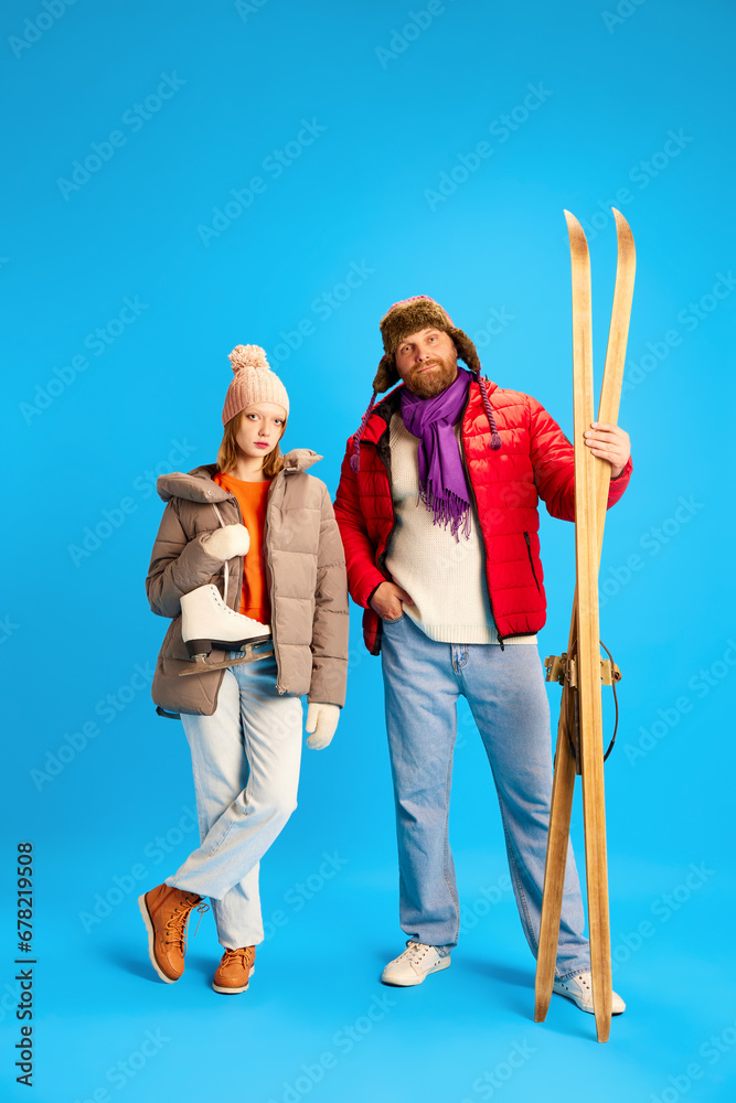 Father and daughter dressed winter warm outfit and holds vintage ski and skates isolated blue studio background. Concept o winter, extreme sport.