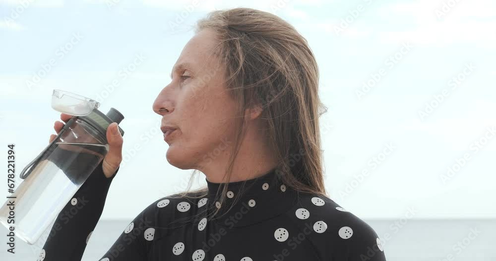 A young woman sits on a sandy beach and drinks water from a transparent plastic bottle against the backdrop of the beach and sea horizon. Profile portrait A young woman sits on a sandy beach and drinks water from a transparent plastic bottle against the backdrop of the beach and sea horizon. Profile portrait