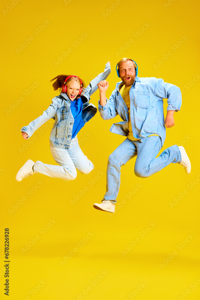 Father and daughter, parent and kid dressed style denim outfit and have fun listening music in headphones and jumping of joy isolated yellow background.