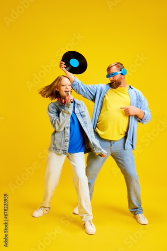 Father and daughter, parent and teenager in style denim outfit have fun with retro music plate and sing to microphone isolated yellow background.