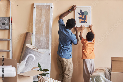 Rear view of African American man and his little son hanging picture on wall of spacious living room during relocation to new apartment photo