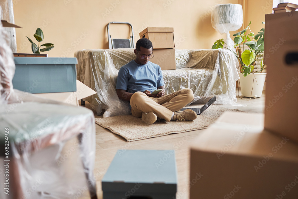 Young African American man with smartphone sitting on the floor by couch covered with cellophane and watching online video Young African American man with smartphone sitting on the floor by couch covered with cellophane and watching online video