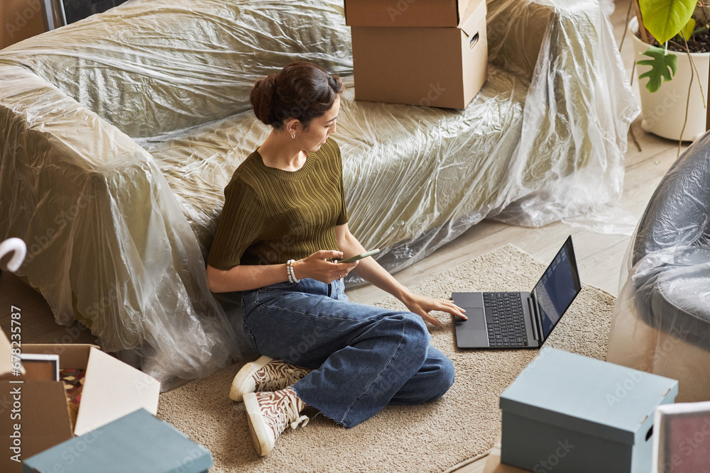 © Seventyfour - Young serious woman in casualwear looking at laptop screen while sitting on the floor by couch in spacious living room of new apartment
