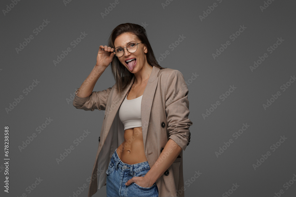 Smiling, chic lady in athletic bra and jacket radiates happiness on a gray backdrop - obrazy, fototapety, plakaty 