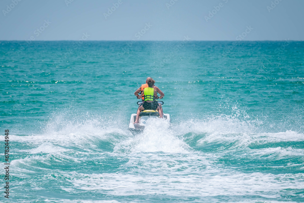 Two persons wearing safety life jackets drive jetski from shore to open sea, splashing water. Tropical beach resort. Close up photo Two persons wearing safety life jackets drive jetski from shore to open sea, splashing water. Tropical beach resort. Close up photo