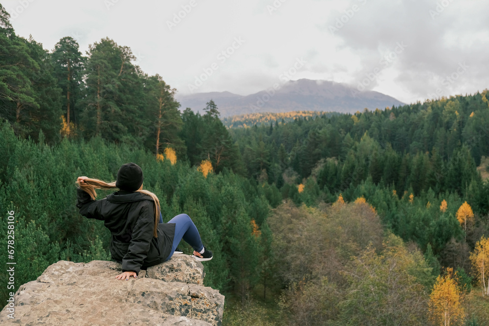 the girl is sitting on a big stone holding her hair in her hands and enjoying the view of the mountains and the autumn forest
