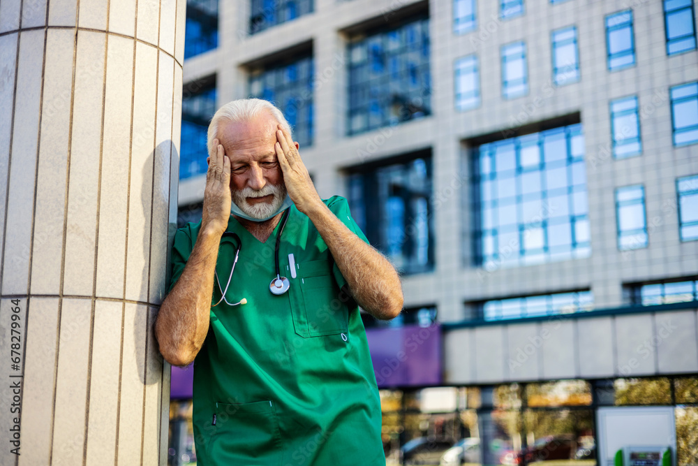 Fototapeta premium Tired exhausted Caucasian male doctor wears green uniform standing in front of hospital.