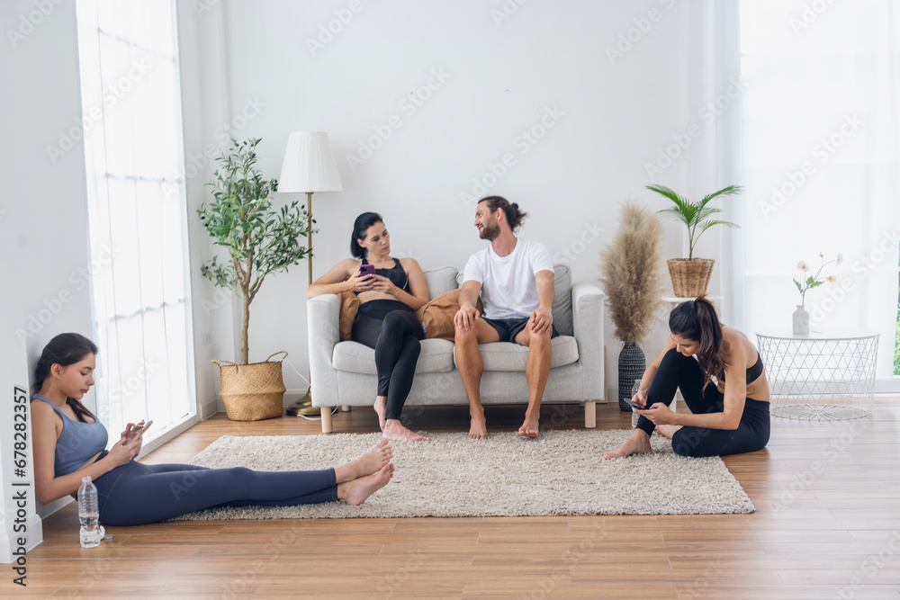 Group of male and female friends with yoga instructor wearing sports clothes, all sitting and holding smartphones after break, exercising, doing yoga class in living room at studio.