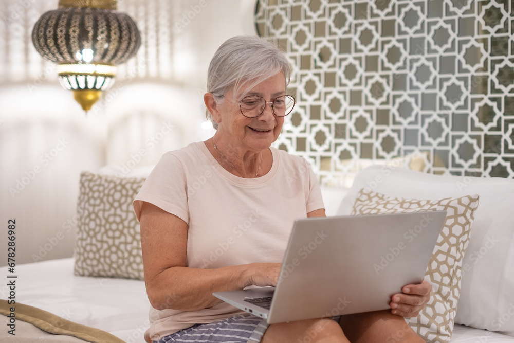 Smiling senior modern woman sitting on bed surfing the net with laptop, white-haired grandmother enjoying technology Smiling senior modern woman sitting on bed surfing the net with laptop, white-haired grandmother enjoying technology