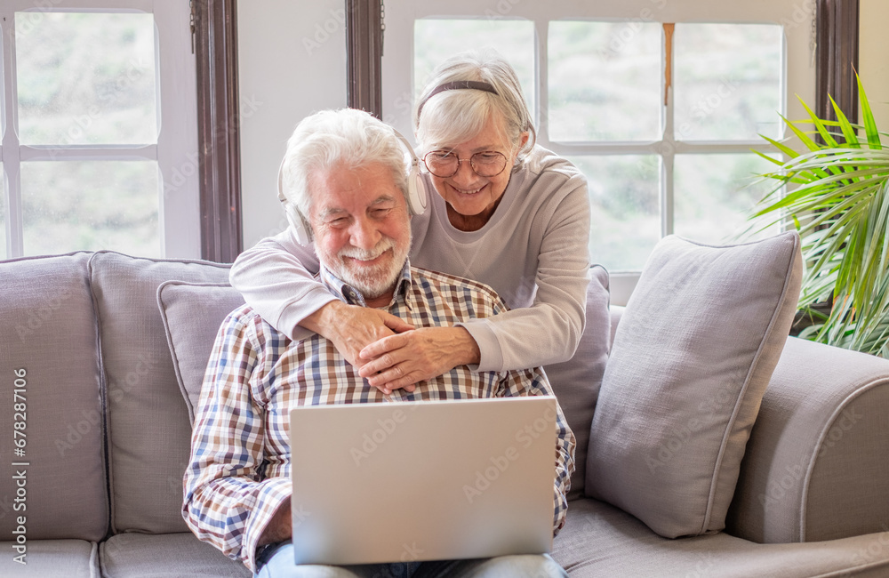 Happy 70s white-haired senior couple relaxing on home couch, looking in internet store using laptop, shopping together online. Old generation and wireless technology users Happy 70s white-haired senior couple relaxing on home couch, looking in internet store using laptop, shopping together online. Old generation and wireless technology users