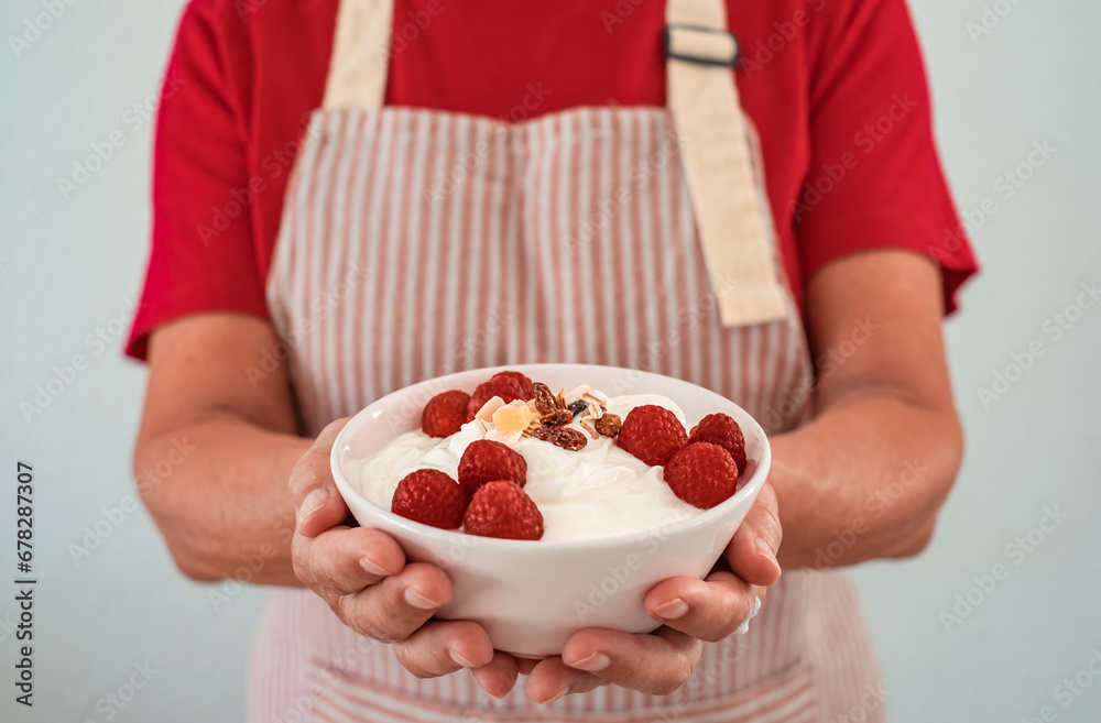 Elderly woman in apron holding bowl of yogurt filled with fresh raspberries and dried fruit. Breakfast or lunch, healthy eating and dietary concept Elderly woman in apron holding bowl of yogurt filled with fresh raspberries and dried fruit. Breakfast or lunch, healthy eating and dietary concept