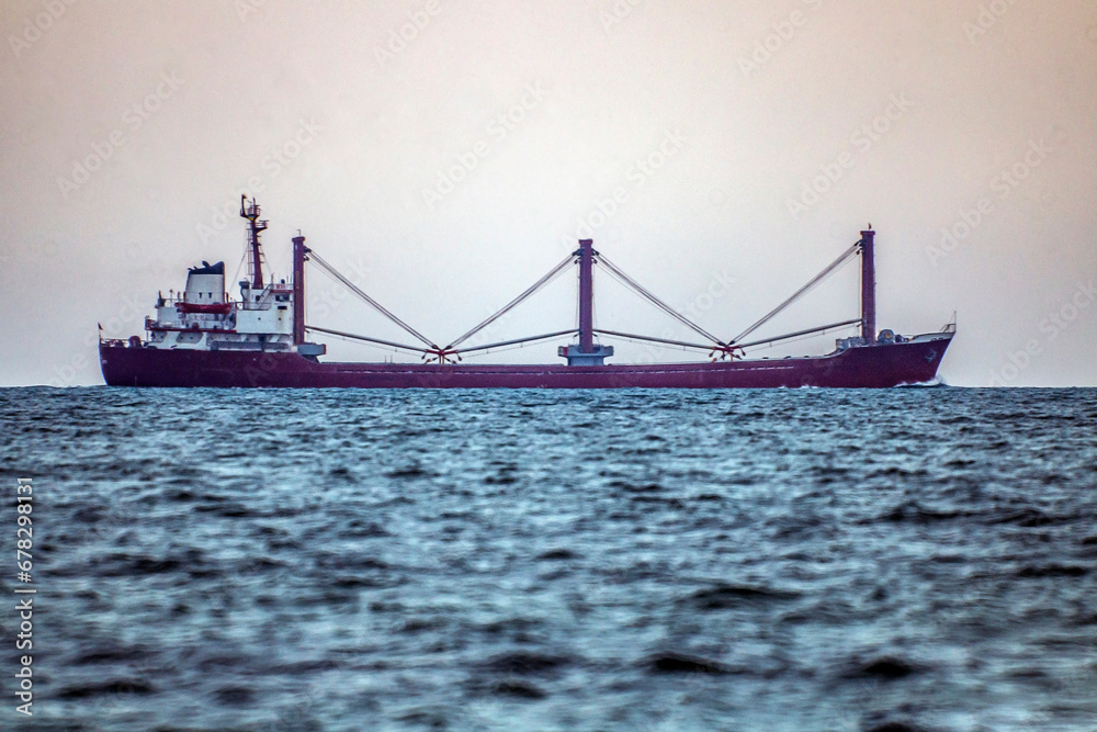 A long red ship with tall cranes on board sails in the ocean on the horizon