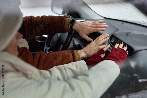 Hands of young couple in winterwear sitting in front of stove inside car during travel and keeping their hands against warm air