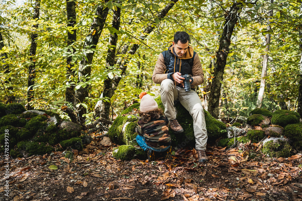 Photographer father with backpack and his fashionable son with hat resting sitting on a stone in the forest after walking on the path