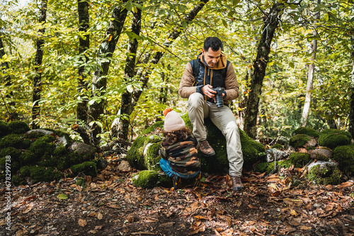 Photographer father with backpack and his fashionable son with hat resting sitting on a stone in the forest after walking on the path
