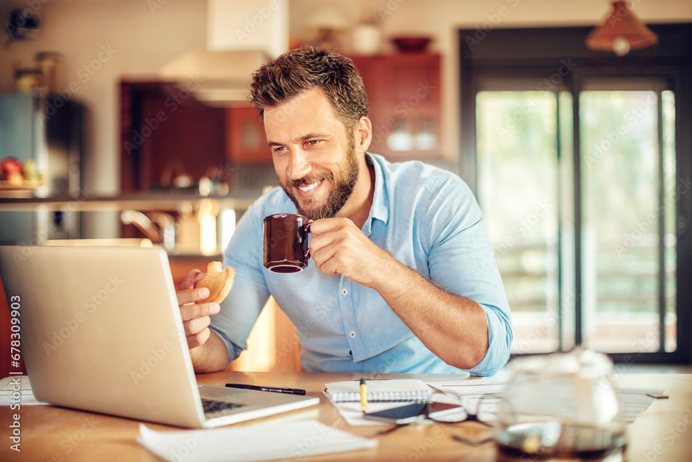Happy young man holding coffee cup looking at laptop in home setting Happy young man holding coffee cup looking at laptop in home setting