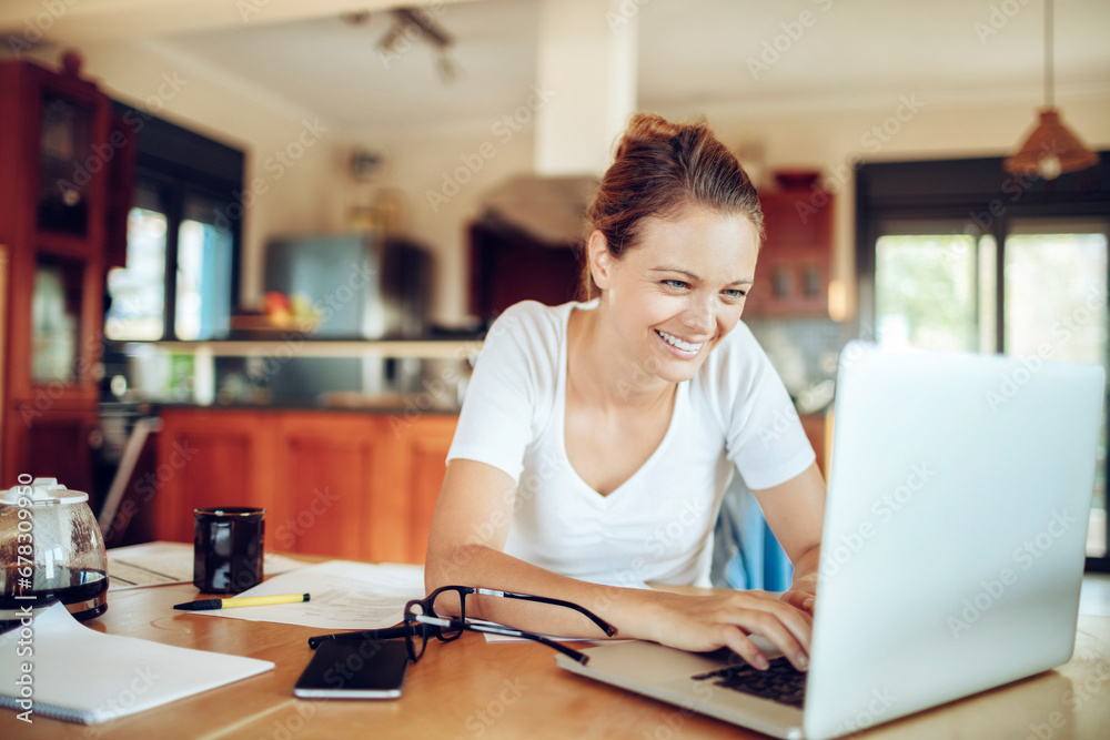 Smiling young woman using laptop with paperwork on desk at home Smiling young woman using laptop with paperwork on desk at home
