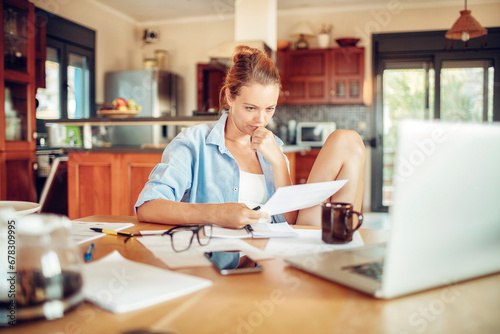 Young woman sitting in home at desk holding document paying bills online with laptop