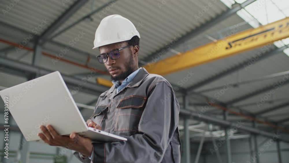 Serious African American engineer using laptop at workplace. Worker wearing helmet and uniform standing in large storage. Inspector of manufacture working with technology device at warehouse.