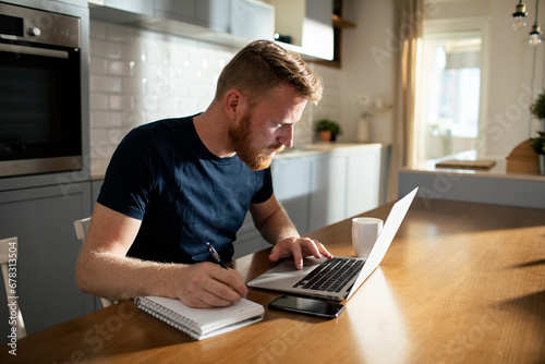 Bearded man using laptop taking notes at home photo