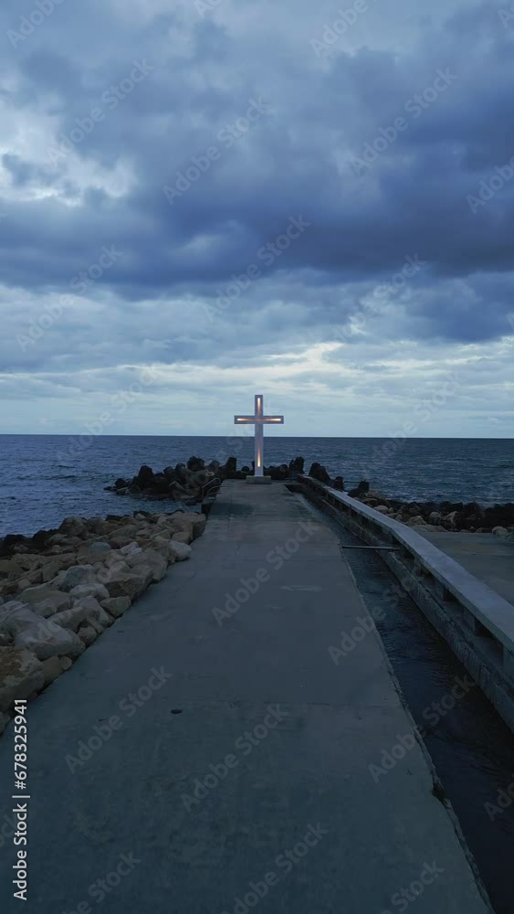 A large Christian cross stands at the edge of a pier against a dramatic sky and sea, seen from above A large Christian cross stands at the edge of a pier against a dramatic sky and sea, seen from above