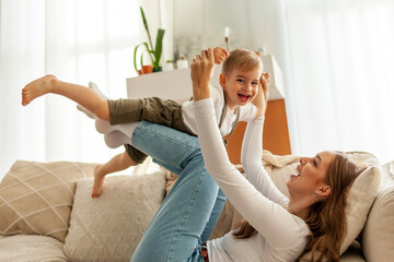 young mother holds and lifts her little son on the sofa, woman plays with her child at home, 2 year old boy flies