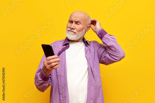 puzzled old bald grandfather in purple shirt uses smartphone on yellow isolated background, confused old pensioner photo
