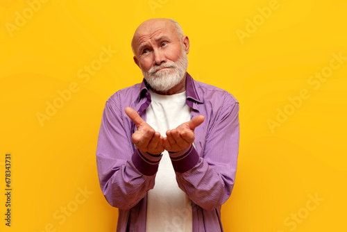 offended bald grandfather in purple shirt begs on yellow isolated background, sad old man pensioner holds empty hands photo