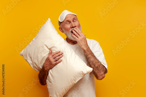 sleepy old bald grandfather in pajamas and sleep mask hugs pillow and yawns on yellow isolated background photo