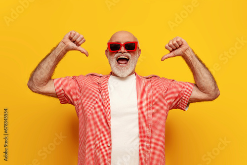 joyful old grandfather in summer shirt and sunglasses points with his hands at himself on yellow isolated background