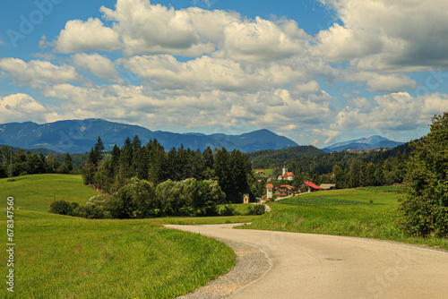 Weg, welcher in eine schöne Landschaft mit kleinem Dorf führt, im Sommer mit Himmel und Wolken