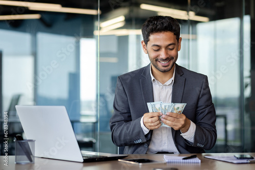 Happy Arab worker in office sitting in front of laptop contentedly counting dollars earned online, success concept, big lottery win.