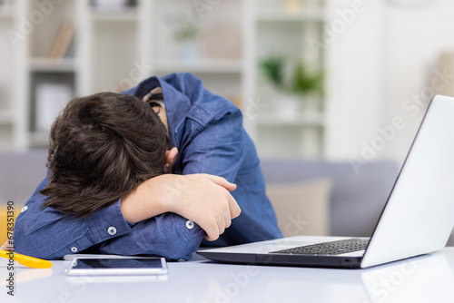 Tired student boy sitting at desk in front of laptop during distance lesson, exhausted sleepy teenager lying on hands on desk with head down, tired of studying.