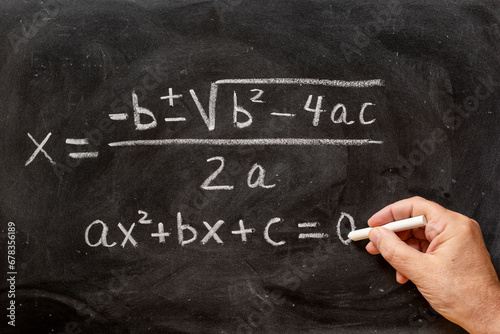 Mano escribiendo ecuaciones cuadráticas en una pizarra con tiza, fórmulas matemáticas