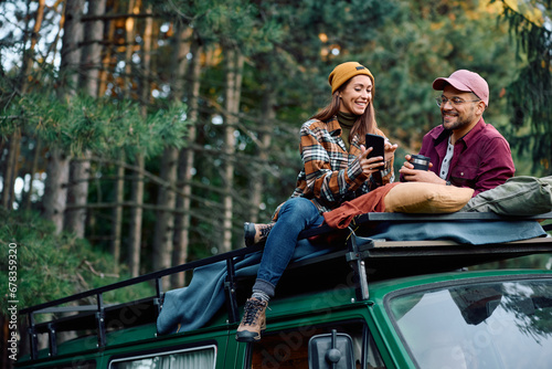 Happy couple using cell phone while sitting on top of their van during camping in woods.